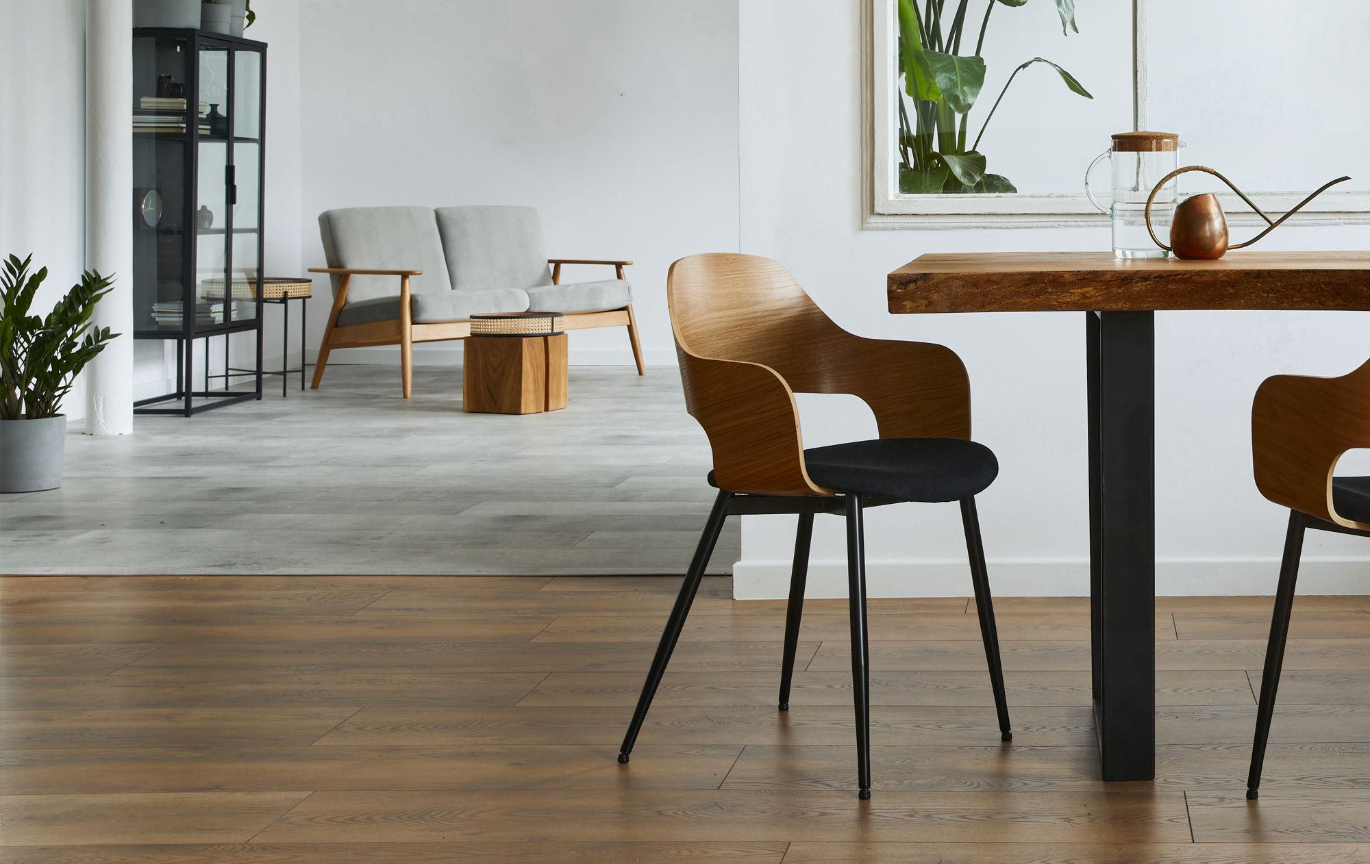 Moderner Essbereich mit einem Holztisch, zwei braunen Stühlen und einer dekorativen Vase. Im Hintergrund sind eine Pflanze, ein Sofa und Regale zu sehen